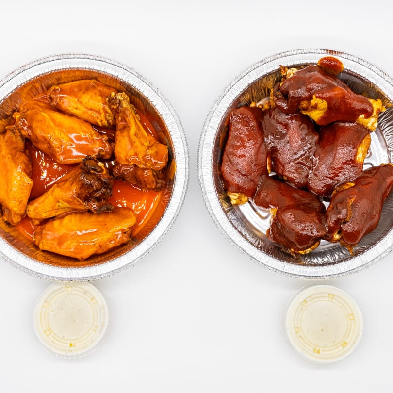 Two foil containers of chicken wings, one with orange buffalo sauce and one with dark BBQ sauce.