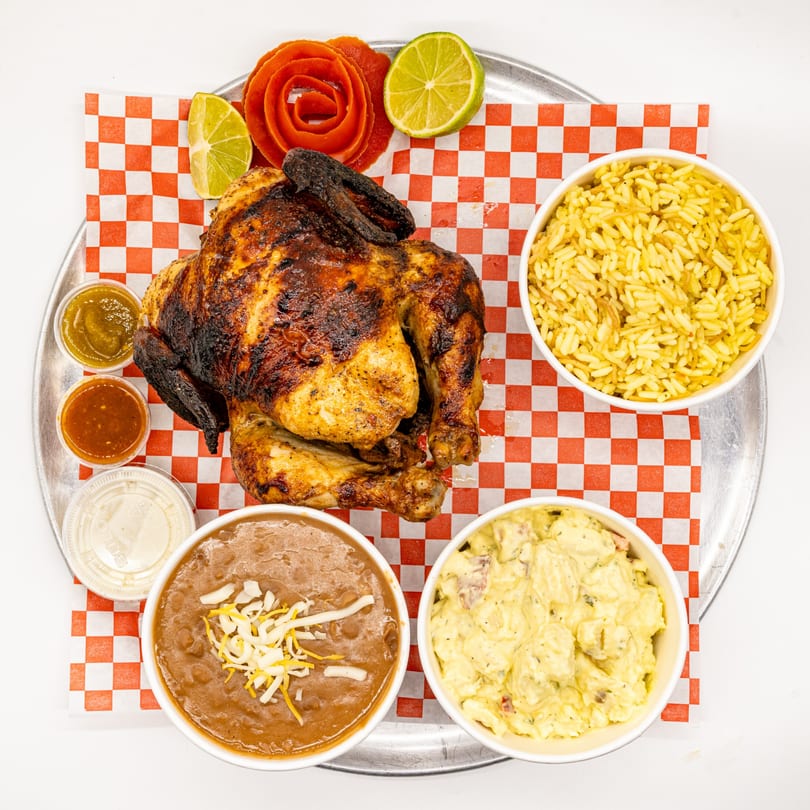Whole roasted chicken with sides of rice, beans, potato salad, and sauces on a metal tray.